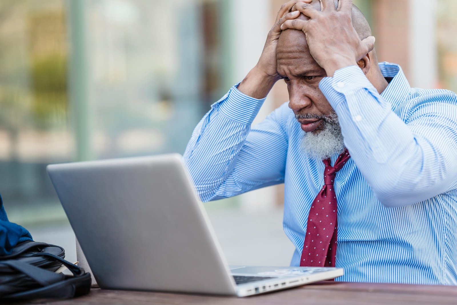 A senior businessman in formal attire looking stressed while using a laptop in an office space.