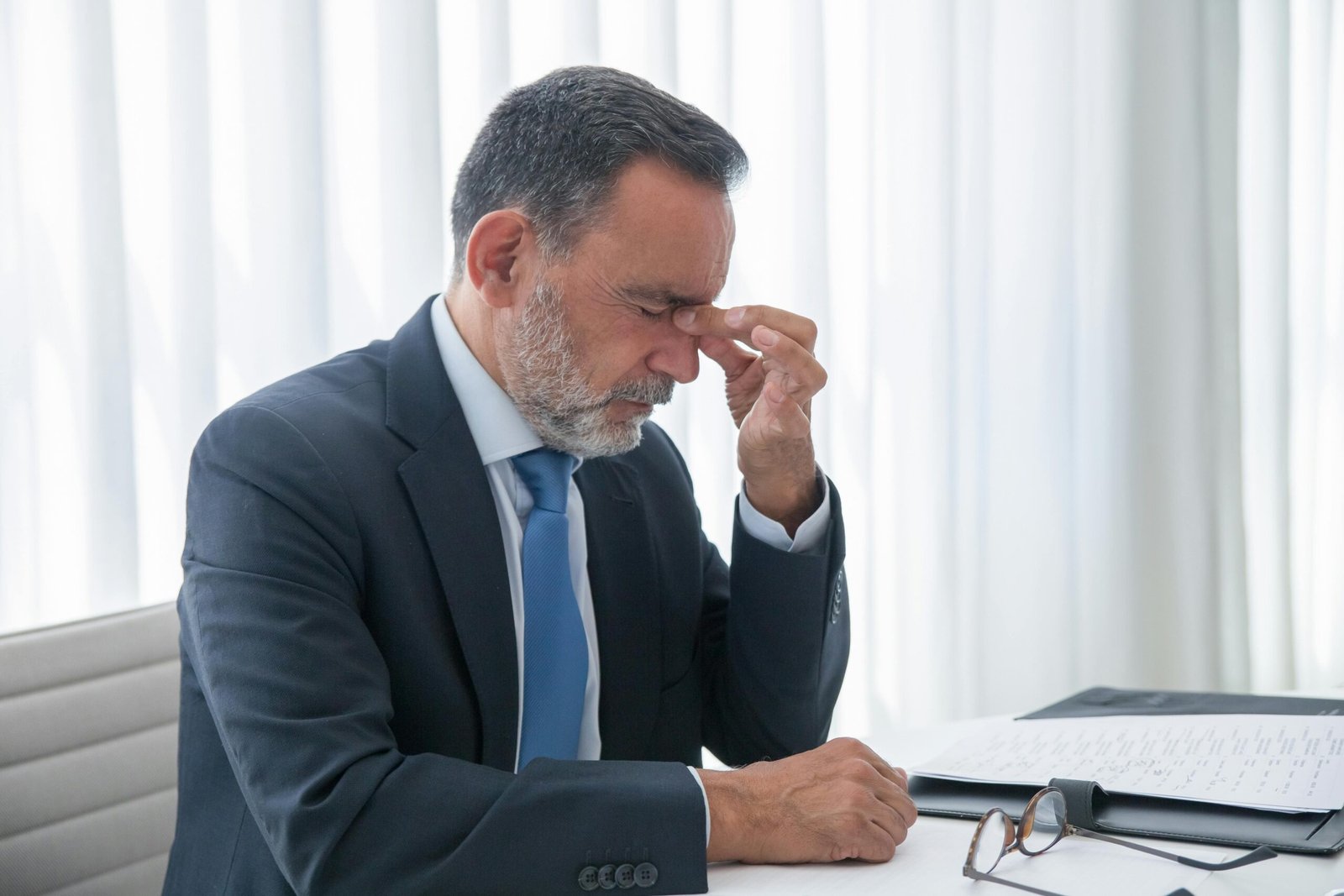 Mature businessman in office, looking exhausted and stressed, with hand on face.